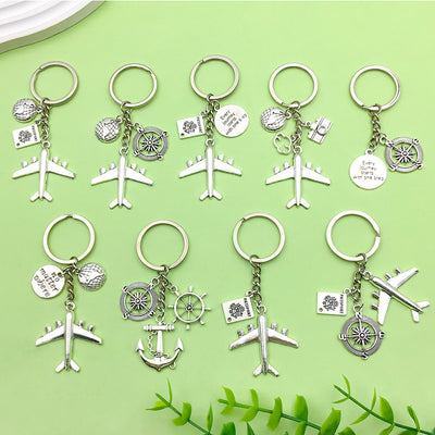 Nine silver-tone travel keychains arranged on green with airplane, globe, compass, passport, camera, quote and nautical 