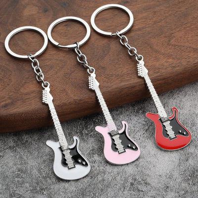 Three electric guitar keychains in white, pink, and red on a wood and gray fabric surface.