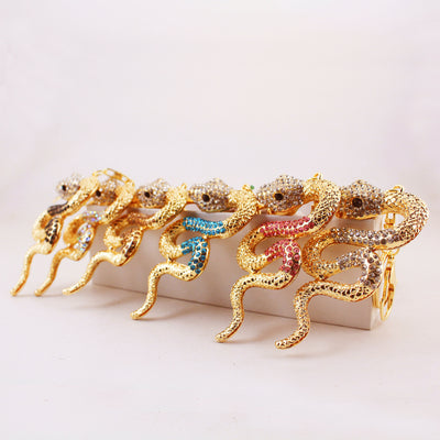 Multiple gold-tone snake keychains with clear, pink, blue, dark, and iridescent stone accents displayed on blocks.