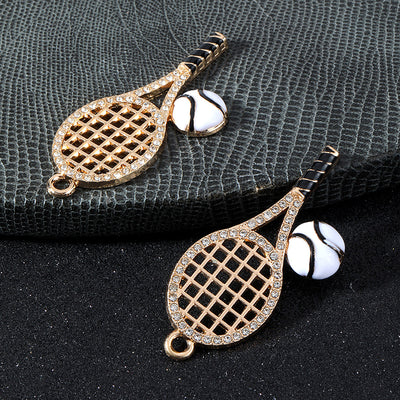 Two gold-tone rhinestone tennis racket charms with black and white enamel tennis balls on a textured grey surface