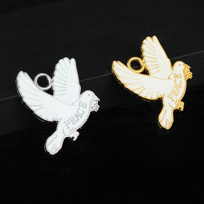 Two white enamel peace dove charms in silver-tone and gold-tone side by side on a dark cloth
