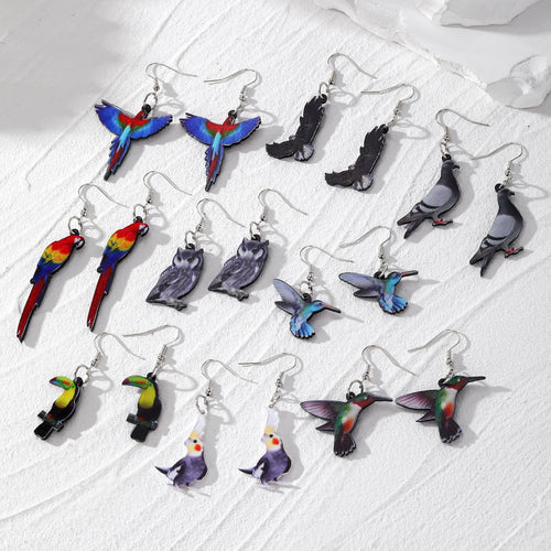 Nine pairs of acrylic printed bird drop earrings laid out on white surface