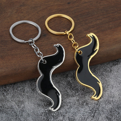 Two black mustache keychains with silver-tone and gold-tone trim on a wood and gray fabric surface.