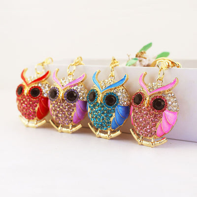 Four gold-tone owl keychains in red, purple, blue, and pink arranged on a white ledge.