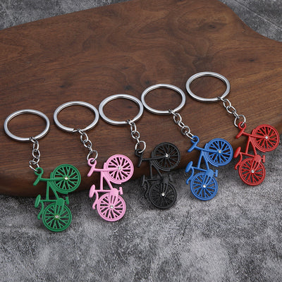 Five bicycle keychains in green, pink, black, blue, and red arranged on wood and gray fabric.