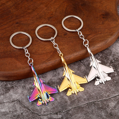 Three fighter jet keychains in iridescent, gold-tone and silver-tone finishes on a wooden surface.