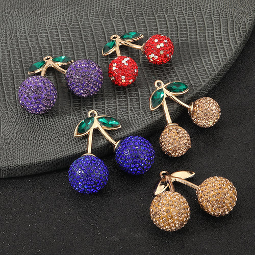 Four pave rhinestone cherry charms in purple, red, champagne and blue arranged on a dark textured surface