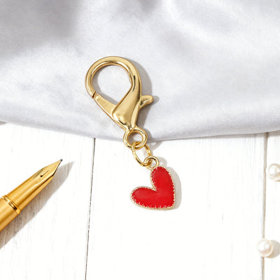 Red heart charm hanging from a gold-tone lobster clasp on white fabric and wood.