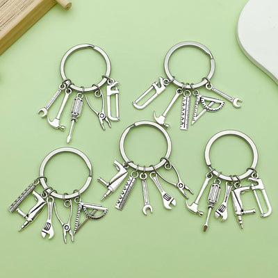 Five silver-tone key rings with assorted miniature tool charms arranged on a green background.