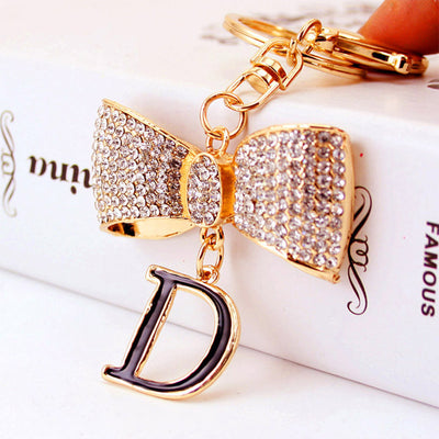 Close-up of a gold-tone keychain with a clear rhinestone bow and black letter D charm.