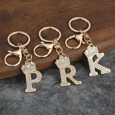 Three gold-tone keychains with crown-topped rhinestone letters P, R and K on a wood and gray surface.