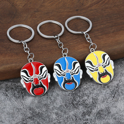 Three Chinese opera face keychains in red, blue, and yellow rest on a wood and gray surface.