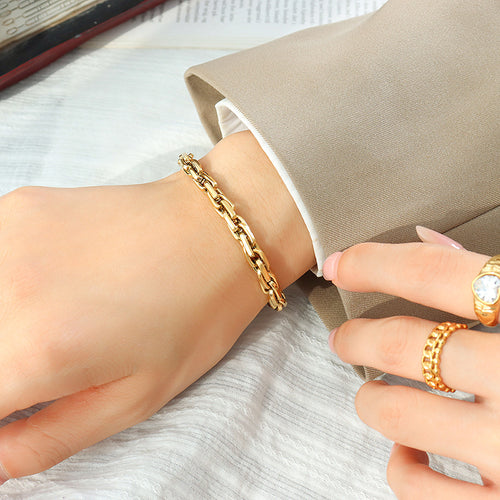 Wrist wearing gold chunky curb link bracelet with beige sleeve and gold rings