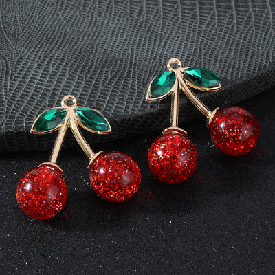 Two gold-tone cherry charms with red glitter cherries and green crystal leaves on a textured gray backdrop