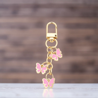 Close-up of a gold-tone keychain with three pink butterfly charms against a blurred neutral background.
