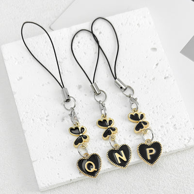 Three black cord phone charm straps with clover charms and heart pendants showing letters Q, N and P on a white tile