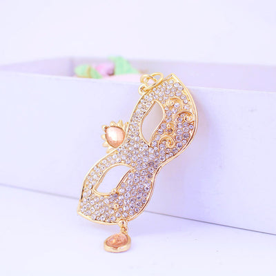 Front close-up of a gold-tone rhinestone masquerade mask against a white box.