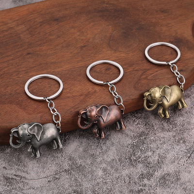 Three elephant keychains in gray, copper, and brass tones displayed on wood and gray fabric.