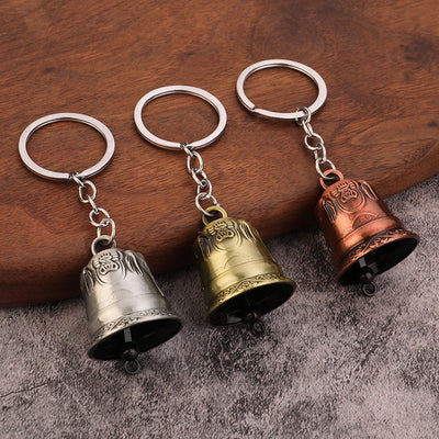 Three embossed bell keychains in brass, copper and silver colors on a wooden surface.