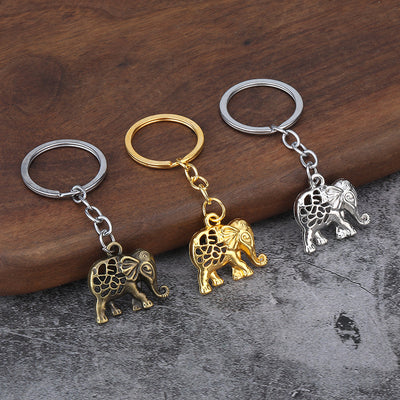 Three elephant keychains in antique bronze, gold tone, and silver tone on a wood and stone surface.
