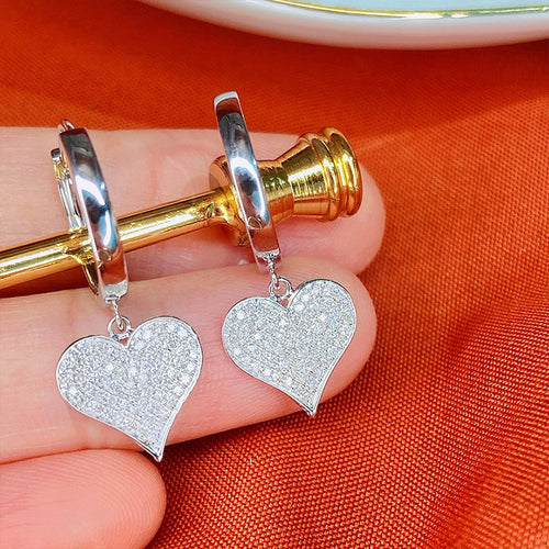 Pair of silver heart charm huggie earrings held between fingers