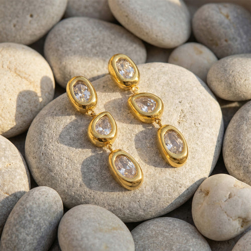 Pair of gold triple zircon drops on gray river stones in sun