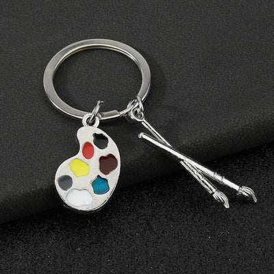 Silver-tone key ring with a seven-color painter's palette and two brush charms on a black background.