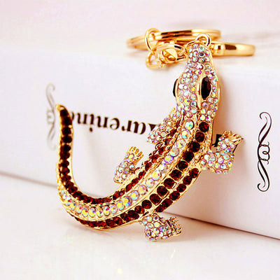 Close-up of a gold-tone rhinestone lizard keychain resting against a white box.