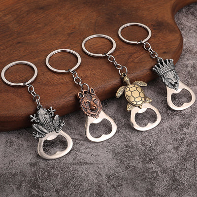 Four metal opener keychains with frog, wolf, turtle and crowned lion motifs on wood and gray fabric.