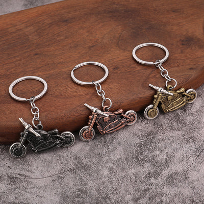 Three motorcycle keychains in dark pewter, copper and bronze finishes on a wooden surface.