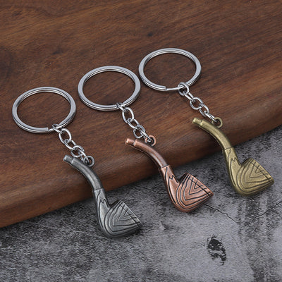Three pipe-shaped metal keychains in silver, copper, and bronze finishes on a wooden surface.