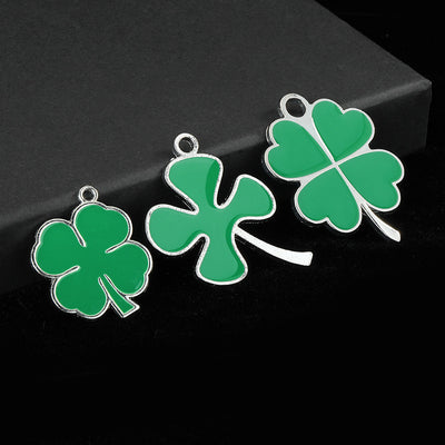 Three green enamel clover charm styles arranged together on a dark box, including a small three-leaf shape