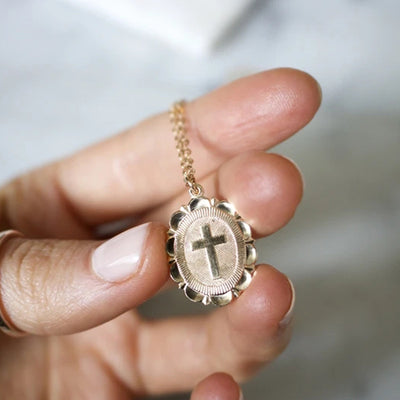 Gold oval scalloped cross pendant held between fingers close-up
