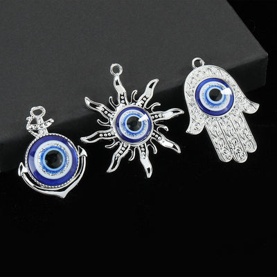 Three silver-tone evil eye charms shaped like an anchor, a sunburst wheel and a hamsa hand with blue glitter resin cente