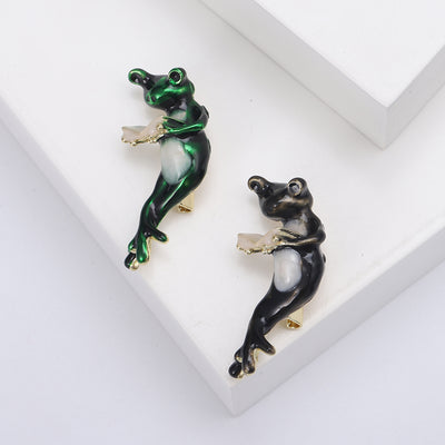 Front view of long legged enamel frog holding an open cream colored book