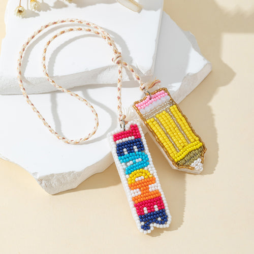 Beaded pencil charm and TEACHER word charm hanging together on a braided cord beside dried flowers