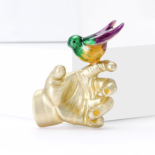 Angled display of small colorful bird perched on a sculpted hand with a pearl-like accent