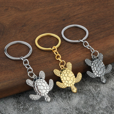 Three turtle keychains in gold, silver, and gunmetal rest on a wooden and gray surface.