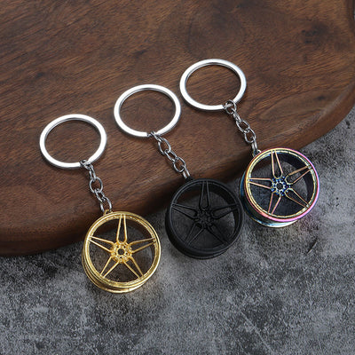 Three five-spoke wheel-shaped key rings in gold-tone, black, and rainbow finishes are arranged on a wood surface.