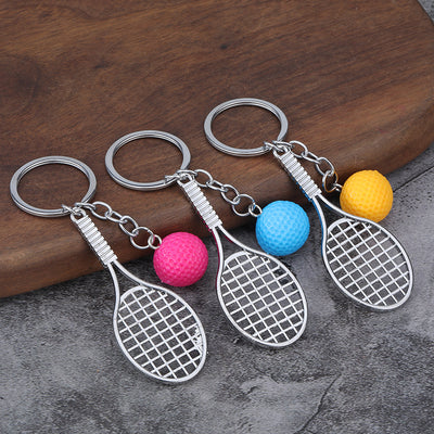 Three silver-tone racket keychains lie beside pink, blue, and yellow textured ball charms.