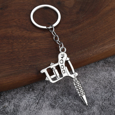 A silver-tone tattoo machine keychain rests on a wood and gray fabric surface.