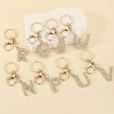 Eight gold-tone rhinestone letter keychains arranged on a cream surface.