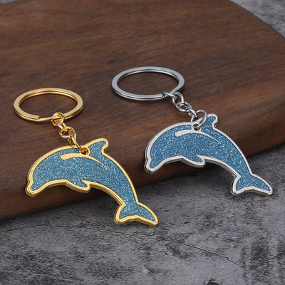 Silver-tone and gold-tone dolphin keychains with blue glitter faces on a wooden surface.