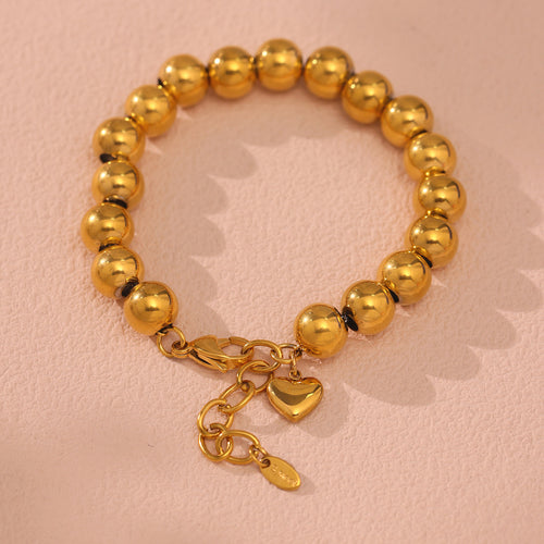 Gold ball bead bracelet with heart charm flat on pink surface