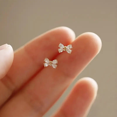 Tiny gold clear CZ bow studs resting on fingertips for scale