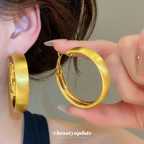 Rounded wide hoops with a smooth brushed gold-tone surface shown in a styled view.