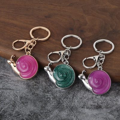 Three snail keychains with pink, green, and purple spiral shells on a wooden surface.