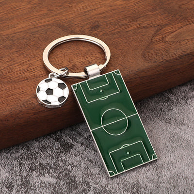 A green soccer field pendant and ball charm rest on wood and gray fabric.