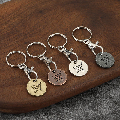 Four shopping cart tag keychains in gold, copper, silver, and dark gray finishes rest on a wooden surface.