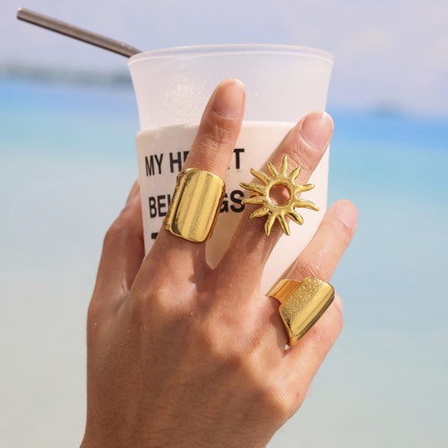 Hand holding iced drink with gold sunburst ring and wide gold bands